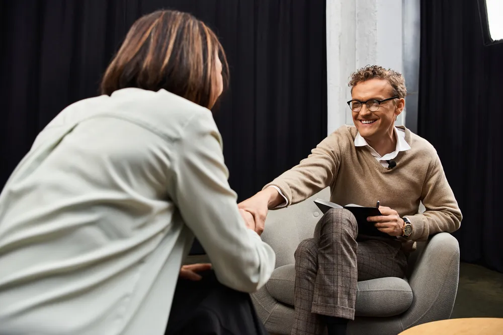 Smiling man, psychologist in English, is interviewed by a journalist on a modern set.