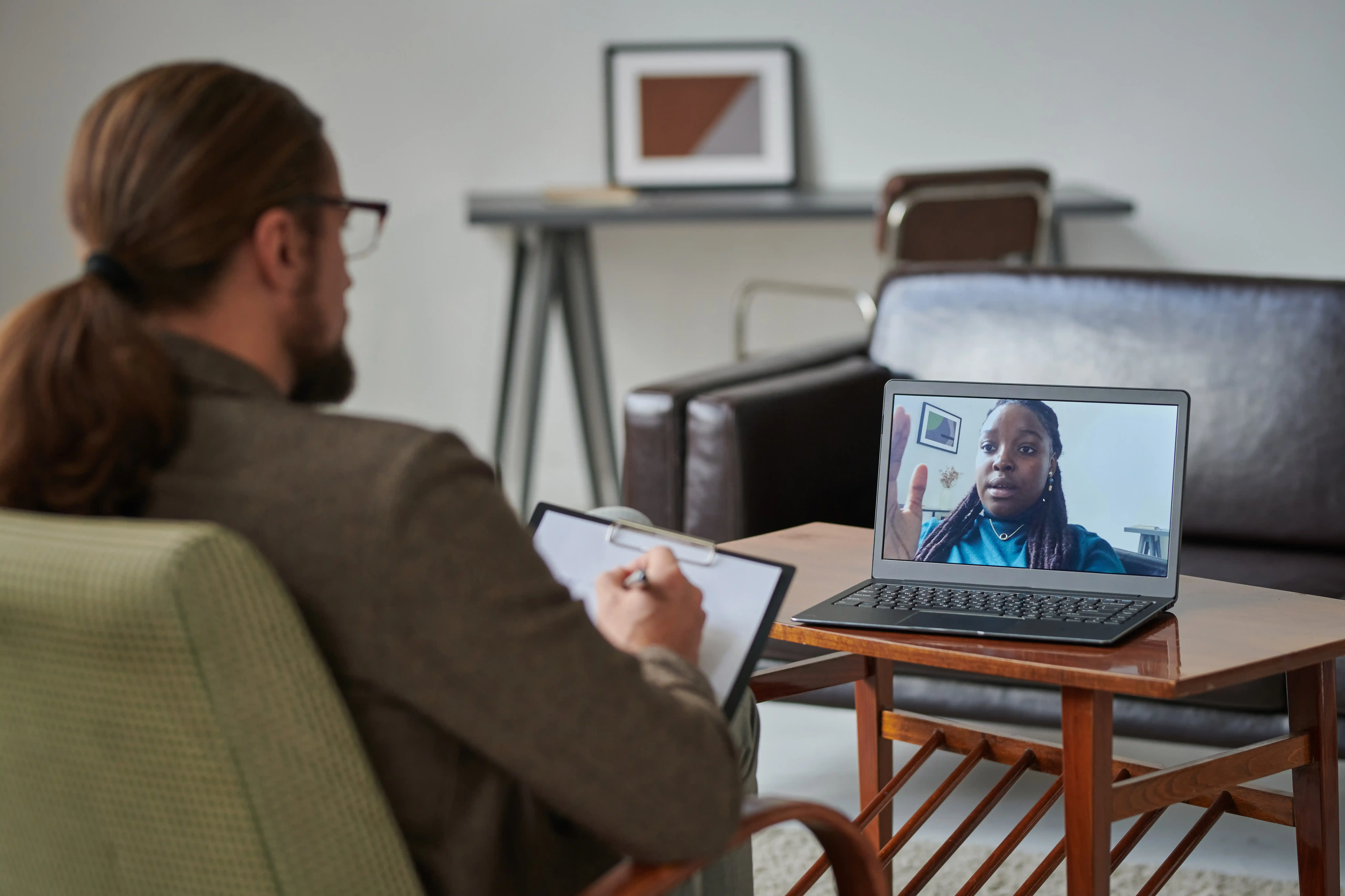 A counselor with glasses takes notes on a table while performing a virtual therapy session with a young patient.