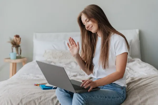 Mujer sentada c&oacute;modamente en su habitaci&oacute;n saludando al iniciar su videollamada de terapia para ansiedad en su computadora.