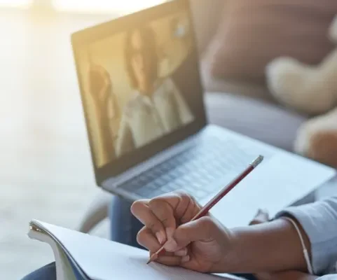 Person taking notes in a notebook during an Acceptance and Commitment Therapy video call on their laptop. Person taking notes in a notebook during an Acceptance and Commitment Therapy video call on their laptop.